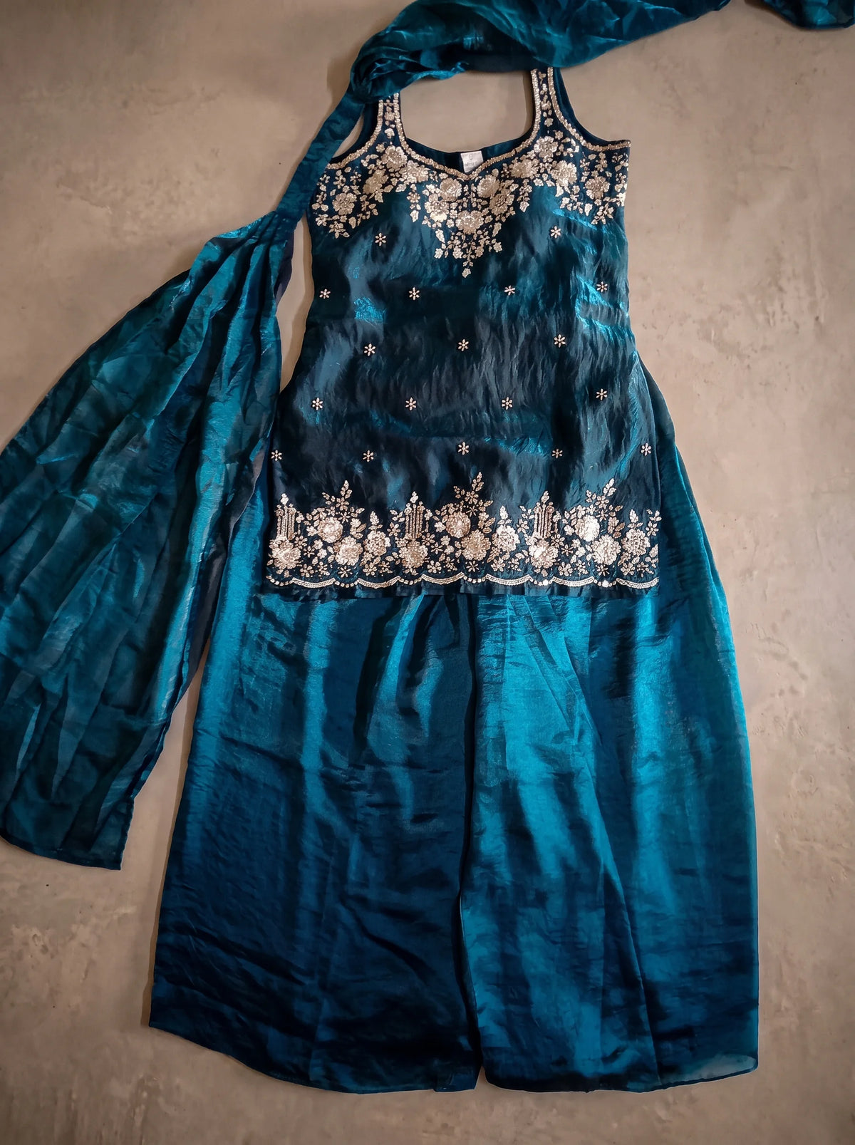 Flat lay of deep teal blue tissue silk sharara set with floral sequin and cut-bead embellished top, flared bottom and matching dupatta.